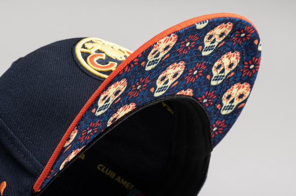 Club America Day of the Dead Snapback Hat with Club America logo embroidered on the front, floral skull embroidered on the side, "Dia de Muertos" embroidered on the back, and marigold and calavera print on the under peak. The hat has a high crown, flat peak brim, and a snapback closure.