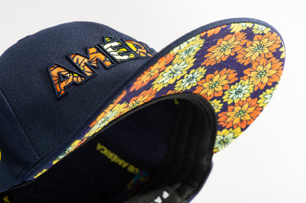 Club America Day of the Dead Snapback Hat with America script with marigold floral overlay embroidered on the front, Club America logo embroidered on the side, "Dia de Muertos" embroidered on the back, and marigold floral print on the under peak. The hat has a high crown, flat peak brim, and a snapback closure.